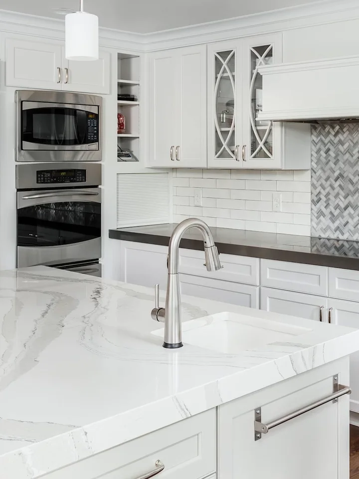 A white kitchen with a marble counter, wall ovens and a tiled splash